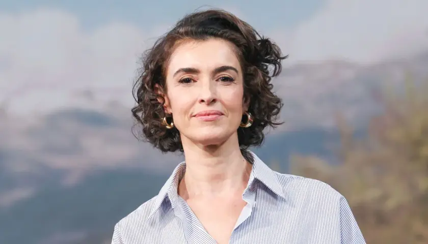 Blanca Romero, con su cabello rizado y pendientes dorados, sonríe con una camisa a rayas.