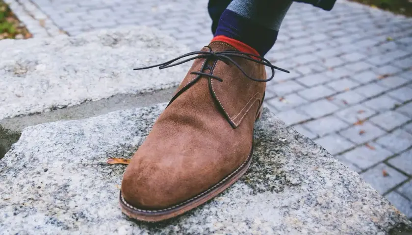 Zapato de gamuza marrón con cordones negros, calcetín a rayas y un consejo sobre como quitar manchas de humedad en zapatos de piel.