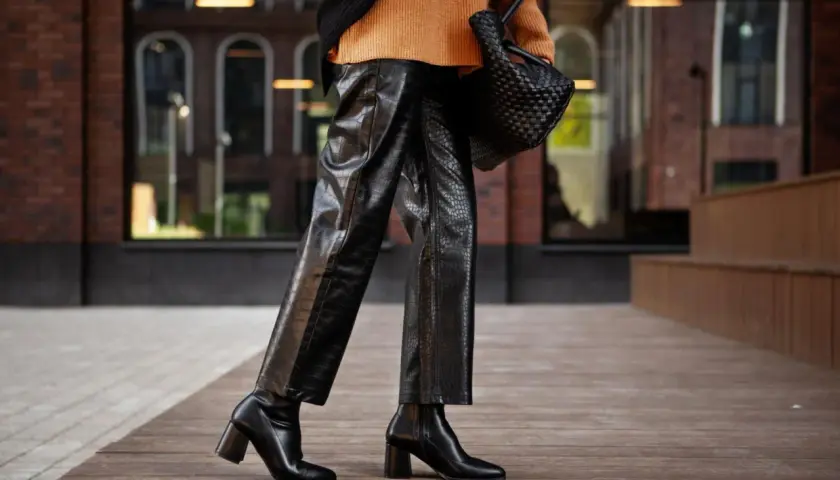 Elegante outfit con botas altas negras, pantalones de cuero texturizado y suéter naranja.