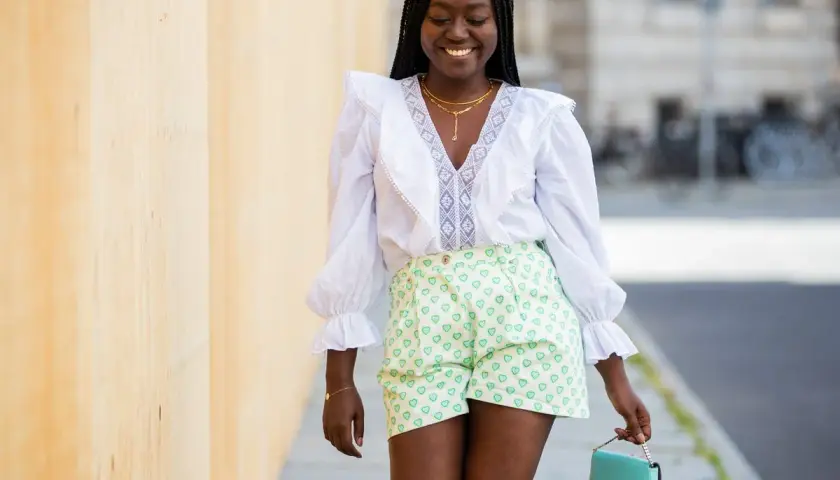 Mujer sonriente con caderas anchas, viste blusa blanca con volantes y shorts verdes con corazones. Lleva un bolso turquesa.