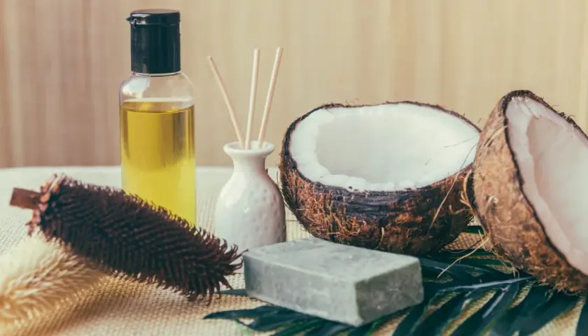 Aceite de coco para el pelo, junto a un coco partido, jabón y un cepillo.