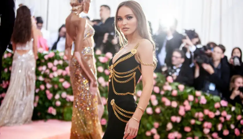 Lily-Rose Depp deslumbrante con un vestido negro adornado con cadenas doradas, posando ante un fondo de flores rosas y fotógrafos.