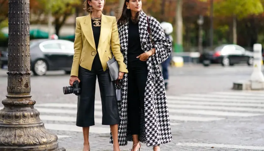 Dos mujeres con estilo muestran cómo vestir en otoño en España: una con blazer amarillo y pantalón culotte de cuero, la otra con abrigo de pata de gallo.