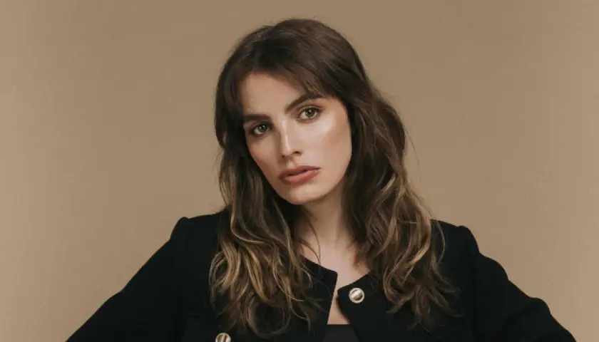 Retrato de una mujer con cabello castaño ondulado y flequillo, luciendo un blazer negro. Su mirada es intensa, transmitiendo un aire de misterio y elegancia, como si fuera la protagonista de una historia de lux pascal.