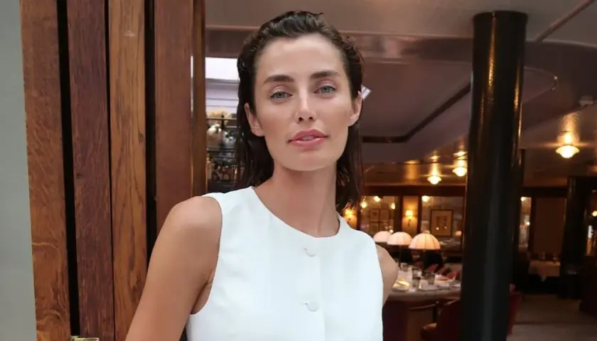 Rebecca Donaldson, con su mirada penetrante y labios rosados, posa con un elegante vestido blanco en un restaurante con ambiente cálido.