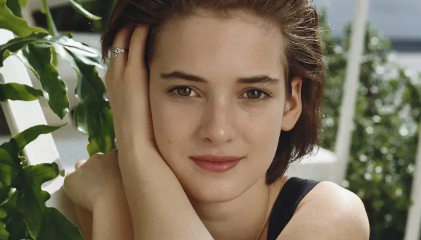 Winona Ryder joven, con su mano en la mejilla y un anillo discreto, rodeada de vegetación exuberante.