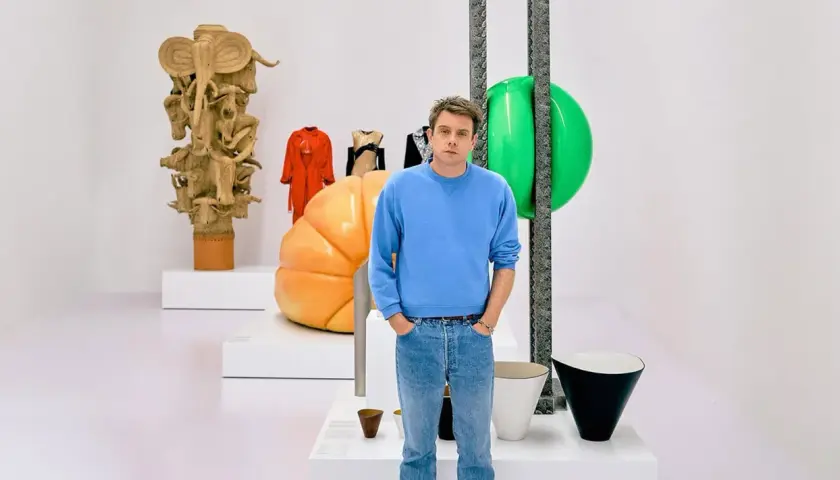 Jonathan Anderson posa entre esculturas y objetos de diseño, incluyendo una figura de madera tallada y un gran globo verde.