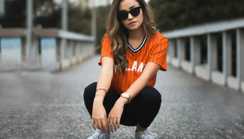Chica con gafas de sol y camiseta naranja a rayas, pantalones negros y zapatillas deportivas. Un ejemplo de estilos de ropa urbana y moderna.