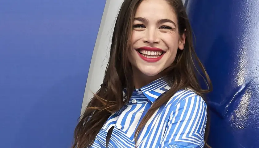 Cosima Ruiz de la Prada, sonriendo con labios rojos y camisa a rayas azules y blancas, se apoya en una pared azul.