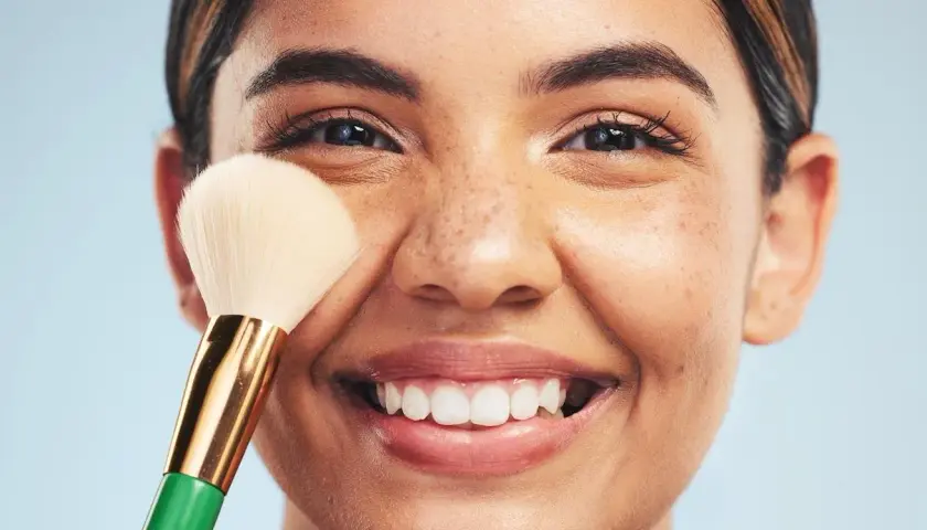 Una mujer sonríe mientras se aplica maquillaje con una brocha. Descubre trucos de maquillaje profesional para realzar tu belleza natural.