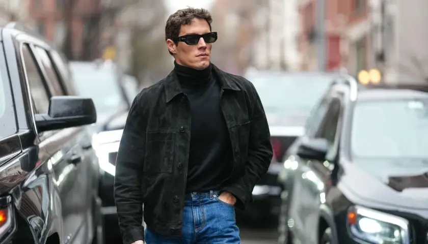 Hombre con estilo casual, viste jersey negro de cuello alto, chaqueta de ante y jeans. Camina por la calle entre coches.