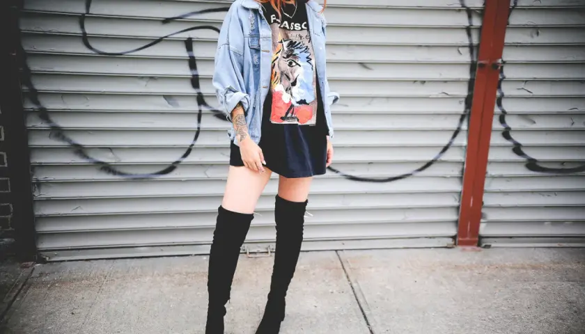 Chica con botas altas para bajitas, chaqueta vaquera y camiseta gráfica, posando frente a una pared de garaje con grafitis.