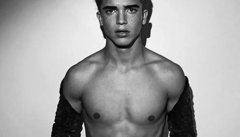 Joven modelo hombre con mirada intensa, torso descubierto y mangas oscuras.