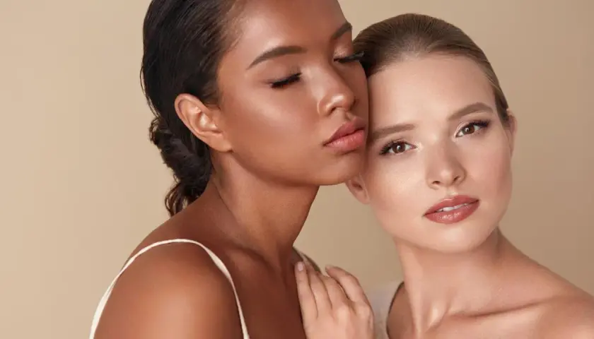 Dos mujeres, una con piel oscura y ojos cerrados, la otra con piel clara y ojos abiertos, comparten un momento íntimo sobre un fondo beige.