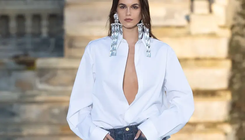 Mujer con camisa blanca desabrochada y vaqueros, luciendo grandes pendientes brillantes.