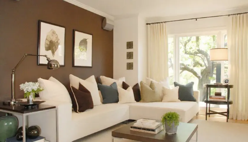 Sofá blanco con cojines en tonos tierra, azul y marrón. La pared marrón crea un ambiente acogedor, mostrando cómo los colores que combinan con marrón pueden ser elegantes.