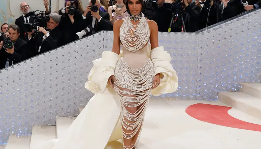 Kim Kardashian desciende la escalera de la Met Gala, deslumbrando con un vestido de perlas y una capa voluminosa, rodeada de fotógrafos.