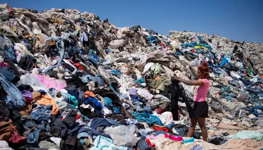 Montaña de ropa desechada, reflejo de las **fast fashion consecuencias**. Una joven busca entre los residuos textiles bajo un cielo azul.