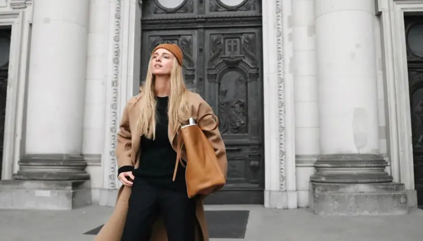 Mujer con abrigo camel, gorro y bolso marrón, luciendo un elegante pantalon negro.