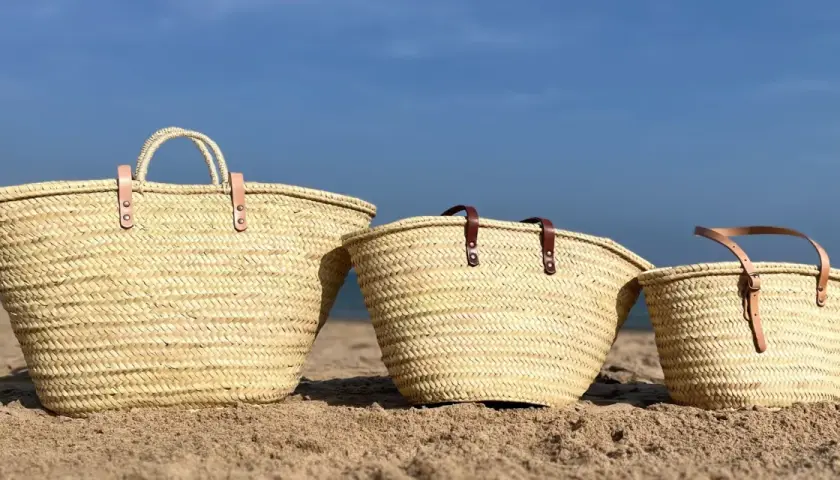 Tres capazos de playa grandes de palma, con asas de cuero, descansan en la arena bajo un cielo azul.