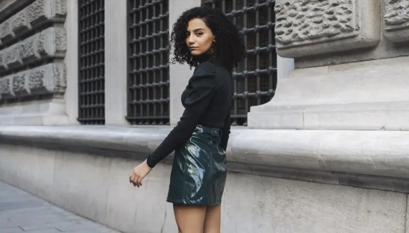 Joven con pelo rizado y falda verde brillante, estilo inspirado en Mary Quant, posa frente a un edificio histórico.