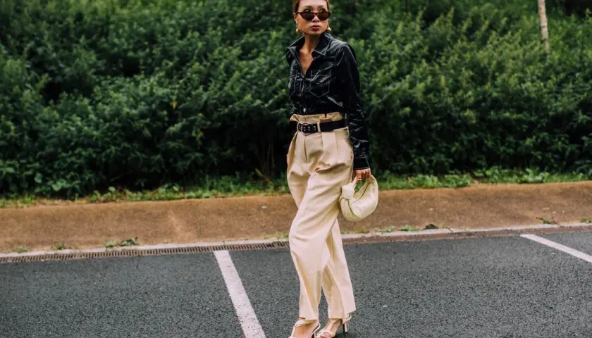 Mujer con elegante **pantalón beige** de talle alto, camisa negra brillante y bolso blanco.