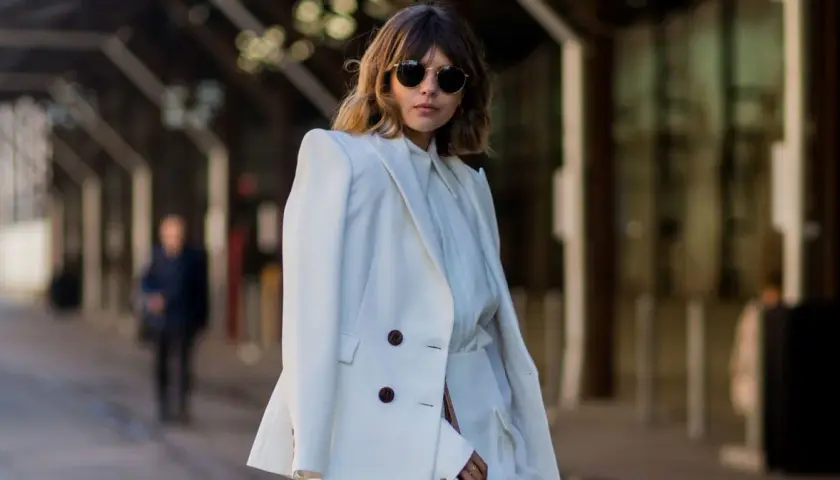 Mujer con traje blanco, gafas de sol redondas y cabello castaño. Su atuendo, elegante y sencillo, refleja el **minimalista significado** de la moda.