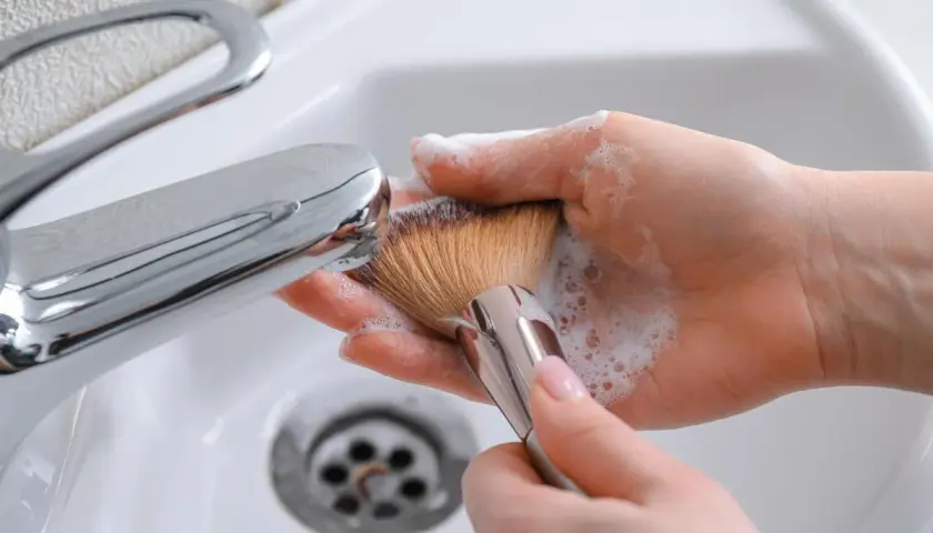 Manos limpiando brochas de maquillaje con espuma y agua bajo un grifo cromado en un lavabo blanco.