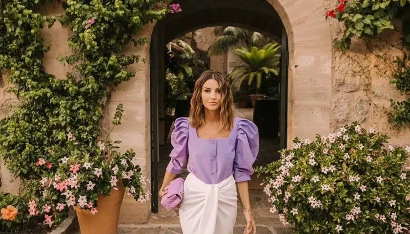 Mujer con ropa que combina color lila claro, un top abullonado y falda blanca, paseando entre vegetación exuberante.