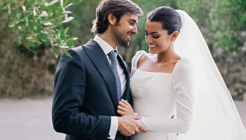 Marta Lozano y su esposo, radiantes en su boda, comparten una mirada cómplice y sonrisas. Él, con traje oscuro y corbata, la abraza tiernamente.
