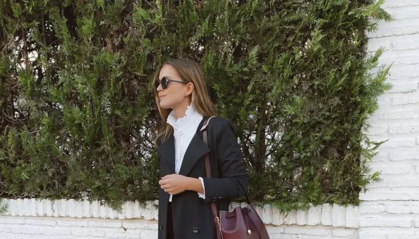 Mujer con elegante outfit para Semana Santa, abrigo negro, blusa blanca con volantes y bolso burdeos.