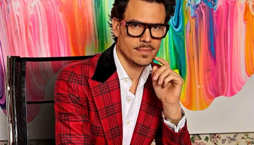 Juan Avellaneda, con su característico traje a cuadros rojos y gafas llamativas, posa con una mano en la barbilla frente a un vibrante cuadro abstracto.