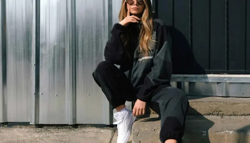Chica con estilo moderno ropa, gafas de sol y zapatillas blancas, sentada en escalones de hormigón frente a una pared de metal.