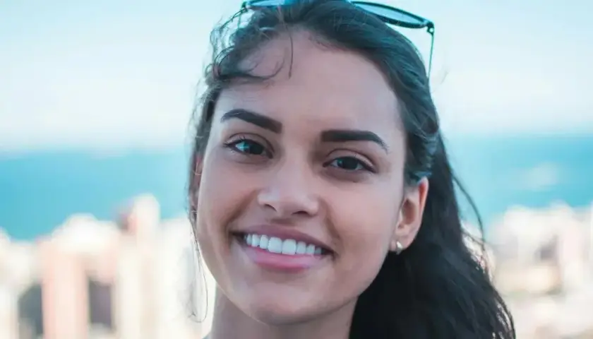 Sonrisa radiante de una joven con rostro ovalado, cabello oscuro y gafas de sol en la cabeza. Fondo de ciudad y mar.