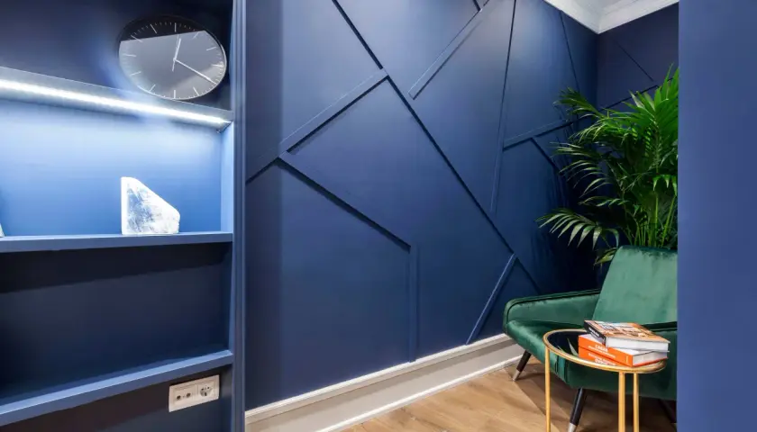 Un sillón verde menta contrasta con la pared azul oscuro. Una planta y libros descansan sobre una mesa dorada.
