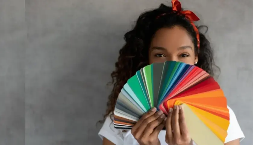 Mujer con cabello rizado y diadema roja sostiene un abanico de muestras de pintura, creando un efecto de color block.