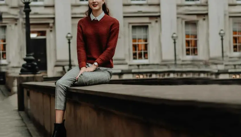 Chica con estilos de ropa modernos: jersey de punto rojo, camisa blanca, pantalones de cuadros y botines negros, sentada frente a un edificio clásico.