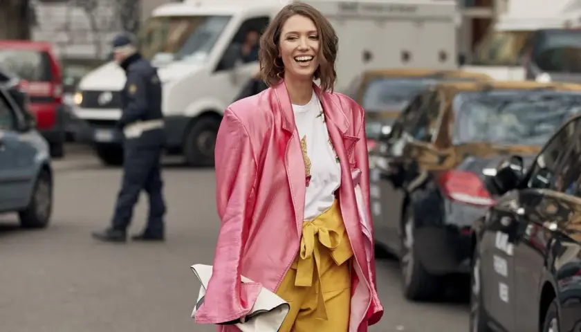 Mujer sonriente con abrigo rosa y pantalones amarillos, demostrando los **colores que combinan con amarillo** en un día urbano.