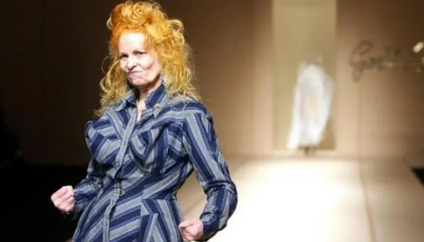 Vivienne Westwood, con su icónico cabello naranja, posa con energía en la pasarela de Westwood, luciendo un vestido a rayas azules y grises.