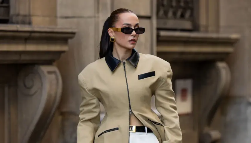 Mujer con gafas de sol oscuras y pendientes dorados, vistiendo una chaqueta beige con cuello negro y cinturón. Un look muy chic.