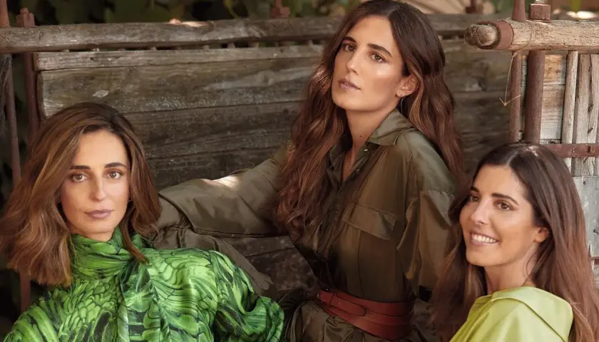 Belen Corsini y dos mujeres más, vestidas con ropa de moda, posan sentadas en un banco de madera rústico.