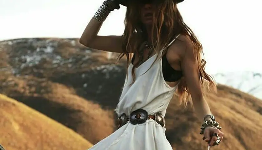 Mujer con sombrero y vestido blanco, luciendo un estilo bohemio con múltiples pulseras y un cinturón adornado.
