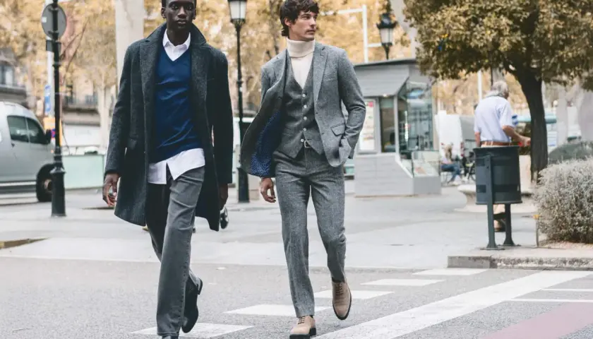 Dos hombres cruzan una calle, uno con abrigo y jersey azul, el otro con traje gris. Representan la moda slow, atemporal y consciente.