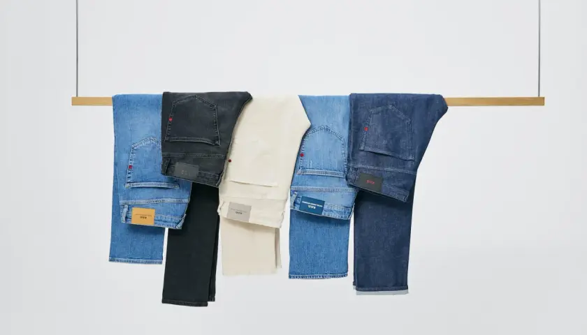 Cinco tipos de pantalones, incluyendo vaqueros azules, negros y beige, colgados en una barra de madera.