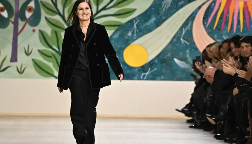 Maria Grazia Chiuri camina en la pasarela con un traje negro, sonriendo ante el público aplaudiendo.
