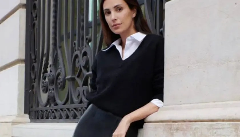 Sassa de Osma, elegante con jersey negro y falda de cuero, apoyada en una columna de piedra junto a una reja ornamental.