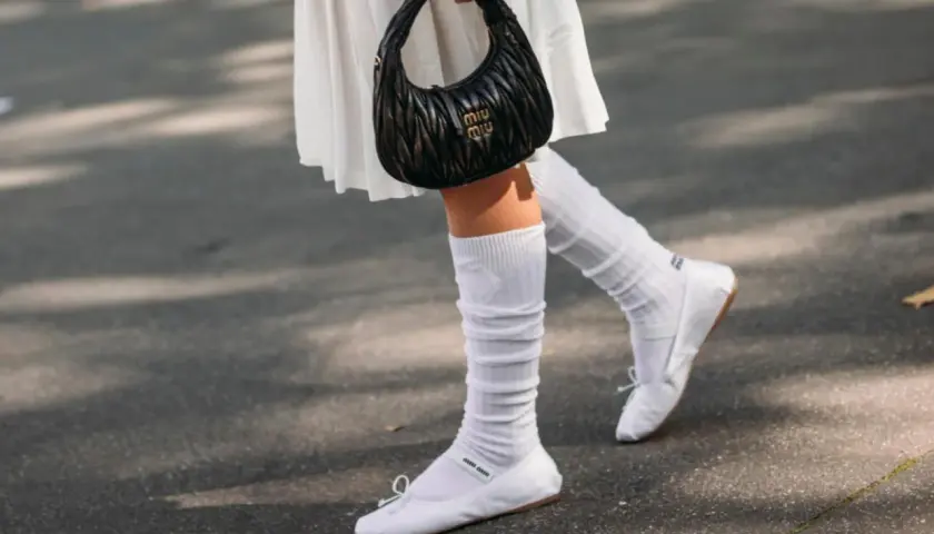 Piernas con bailarinas blancas y calcetines blancos arrugados, un bolso negro Miu Miu en la mano.