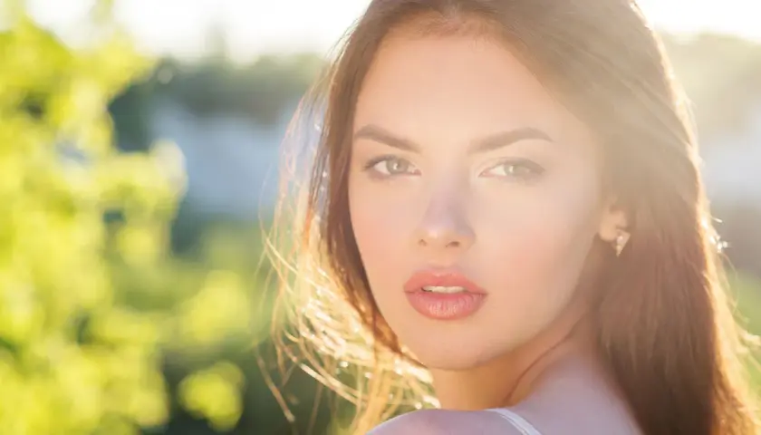 Una joven con un brillo natural, un verdadero glow up, mira a la cámara con el sol iluminando su rostro y el fondo verde desenfocado.
