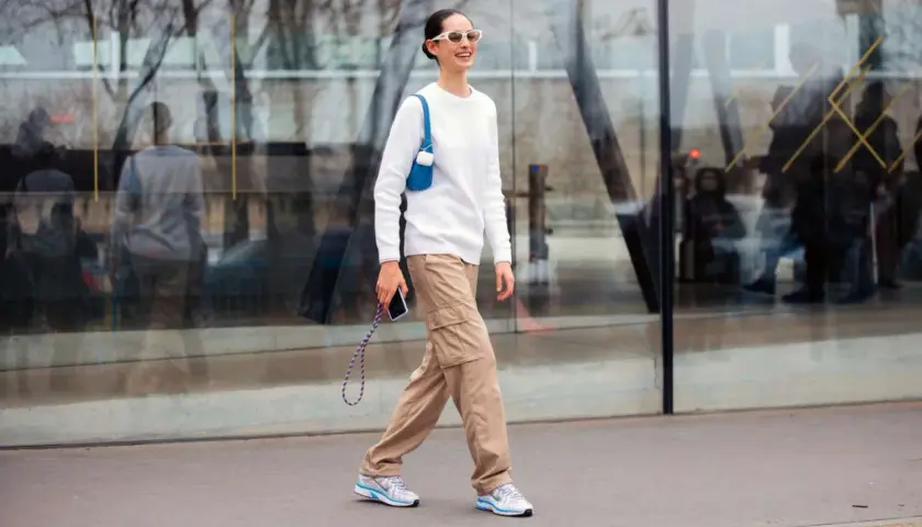 Mujer sonriente camina con un suéter blanco y un pantalón beige de carga, luciendo gafas de sol blancas y un bolso azul.