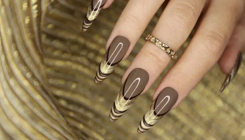 Uñas marrones tendencia con diseño de líneas doradas y brillo. Un anillo dorado adorna el dedo.
